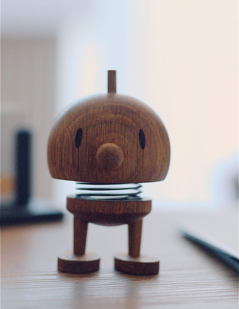 Der Hoptimist Hoptimist - Holzfigur mit einem runden Kopf, Federn und vier Beinen auf einem Tisch.